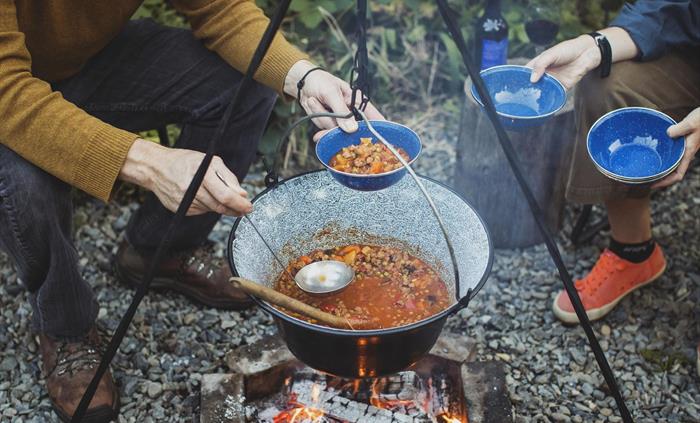 A campfire with people surrounding it. A large pot hangs above full of a steaming stew which is being ladled out into bowls.